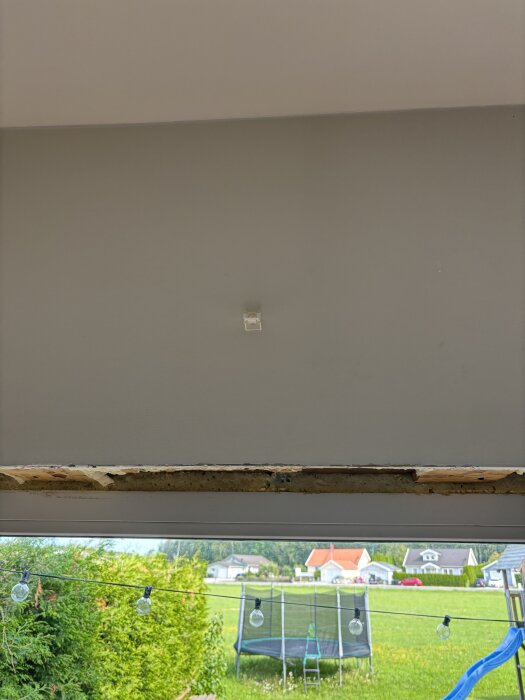 Vy från insidan av huset med en trasig karm, utanför ses en trädgård med studsmatta och hus i bakgrunden.