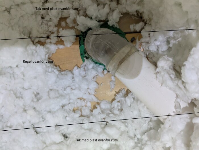 Centraldammsugarrör genom en regel i isolering, med plasttäckta ytor ovanför rum och exponerat trä ovanför vägg. Tejpad öppning vid röret.
