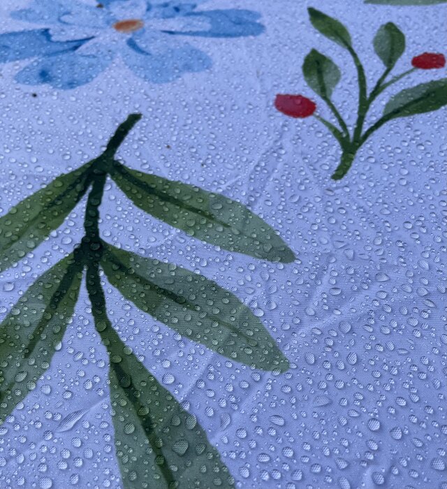 Närbild på blommigt mönster med gröna blad och blå blomma på tyg, täckt av vattendroppar.