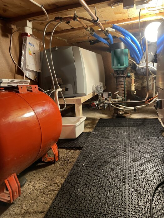 Trångt utrymme med en varmvattenberedare i ett hörn, omgiven av rör och annan VVS-utrustning, samt en röd trycktank på golvet.