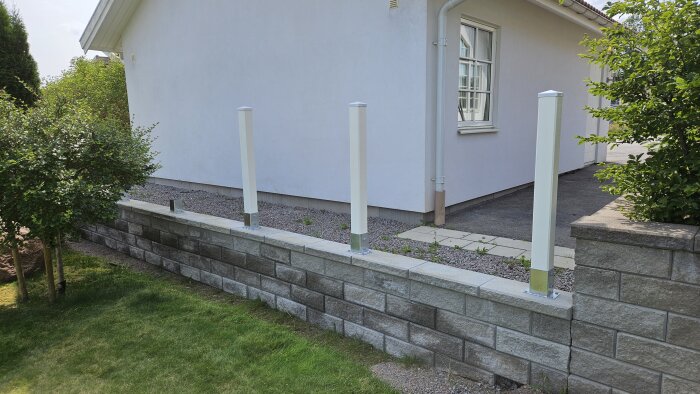 Vit putsad fasad och en låg mur med grönt nedanför. Fyra vita stolpar är monterade längs muren, redo för ett insynsskydd.