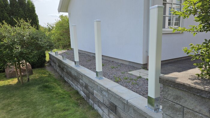 Vit husfasad med grå mur och skruvade stolpfötter redo för staketmontering. Gröna buskar och träd omger området.