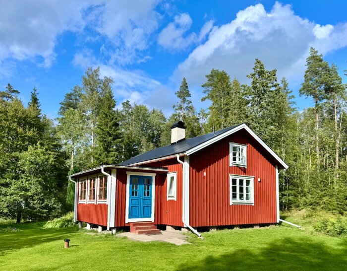 Rött hus med vitmålade detaljer och blå dörrar i S4040-B fönsterfärg, beläget i en skogsmiljö under en klarblå himmel.