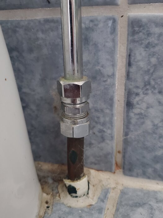 Bilden visar ett kromat vattenrör med en 10 mm anslutning, kopplad till en toalettstol, mot en kaklad vägg.