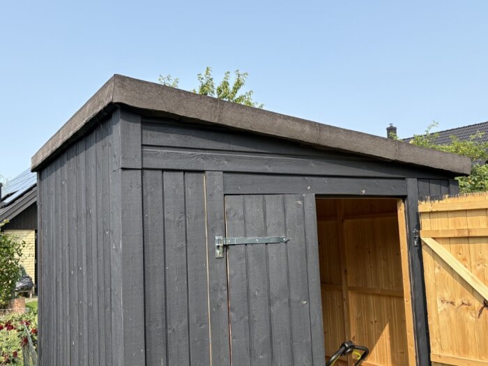 Svartmålat träskjul utomhus med tak täckt av takpapp, dörren är delvis öppen och solen skiner över taket.