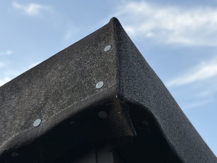 Närbild av ett skjultak med takpapp, där spikar syns längs kanten och på ovansidan mot en blå himmel.