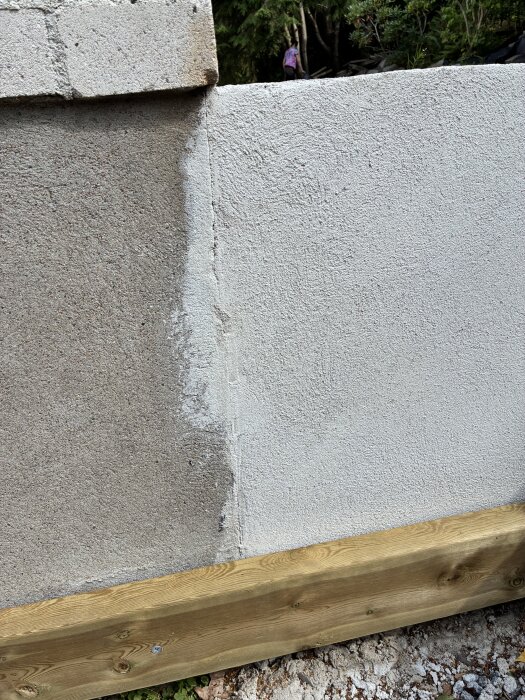 Mur med ljusa fläckar av putsbruk mot en husgrund, bredvid en träplanka.