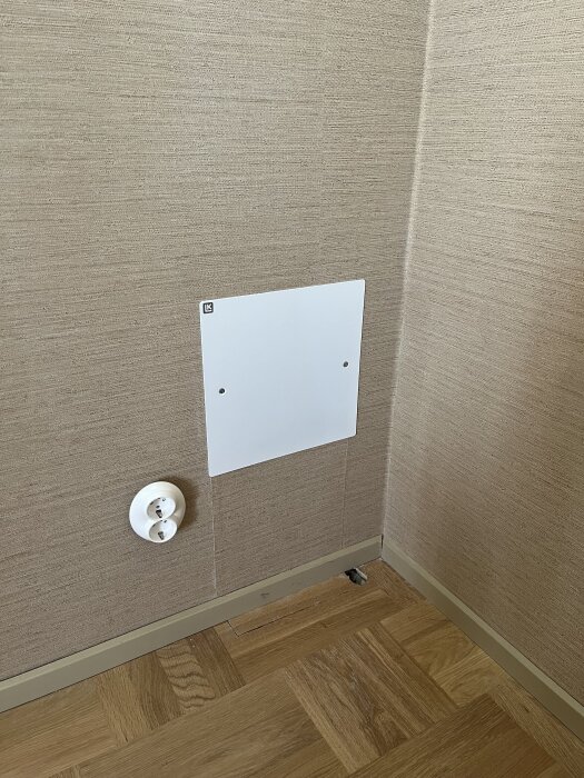Vitt LK-skåp på tapetserad vägg bredvid vägguttag, med parkettgolv som saknar en bit vid sockeln.