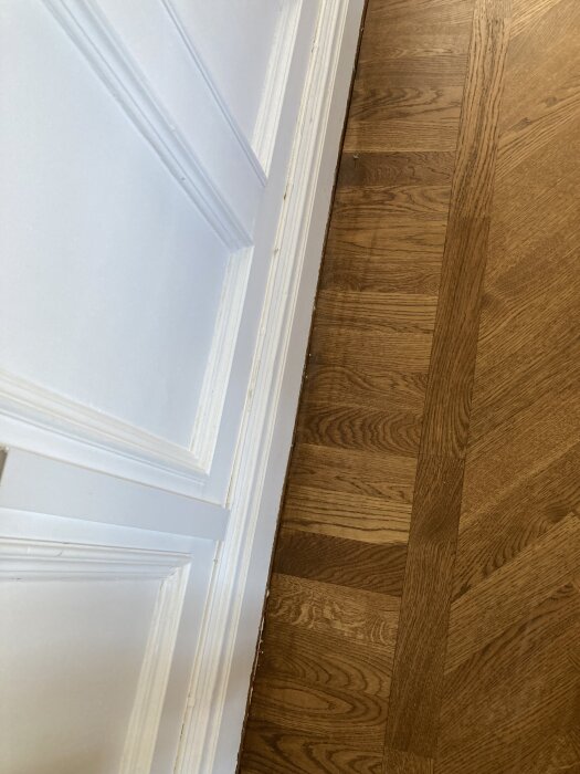 Fiskbensparkettgolv i ek med valnötskulör, synlig nyansvariation längs en vit vägg.