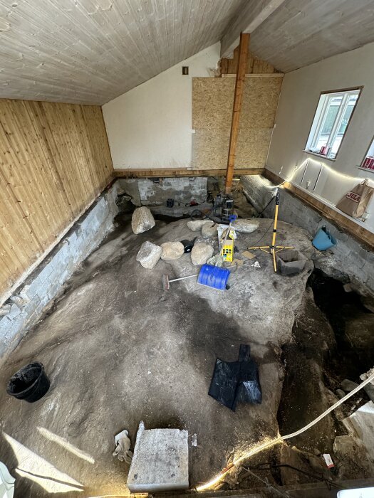 Riven krypgrund med synligt berg, stenar och verktyg, inklusive blå hink och kratta; pågående renoveringsprojekt i en väldig grund.