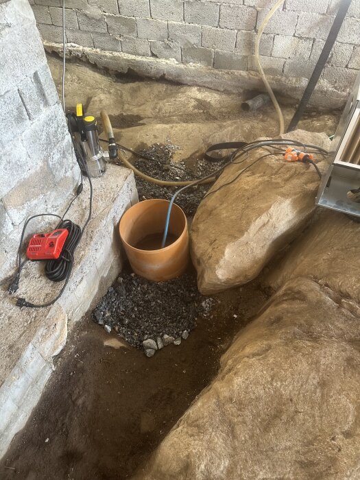 Krypgrund med grävda utgrävningar, ett PVC-rör, en vattenpump och en hink bredvid stora stenar och en slänt av betongblock.