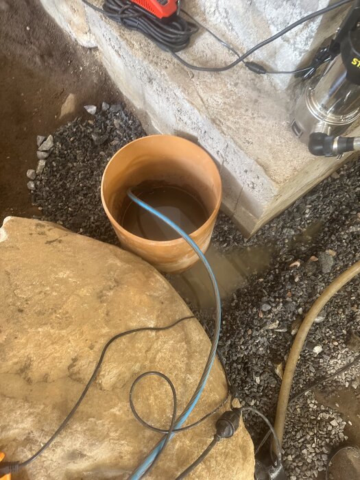 Orange PVC-rör i krypgrund med vatten och kablar runt omkring, stenig mark och del av betongvägg syns.