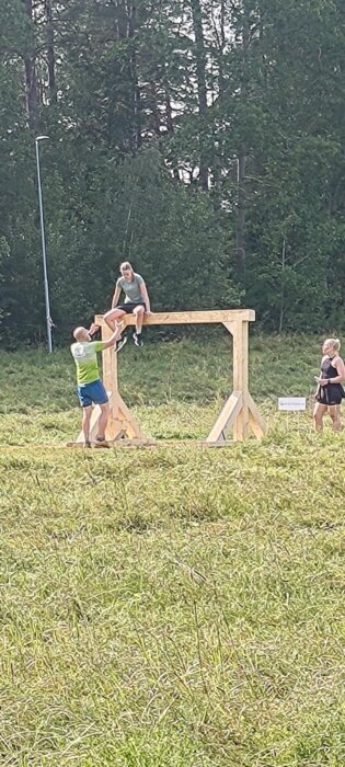 Personer hjälper varandra över ett träöverhäng i en utomhusbana omgiven av skog.