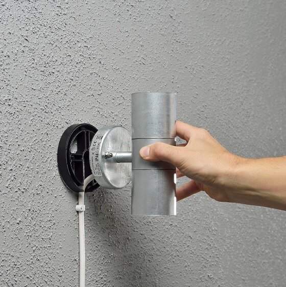 En hand håller i en metallcylinder som är ansluten till en rund väggmonterad platta med sladd, på en grov vägg.