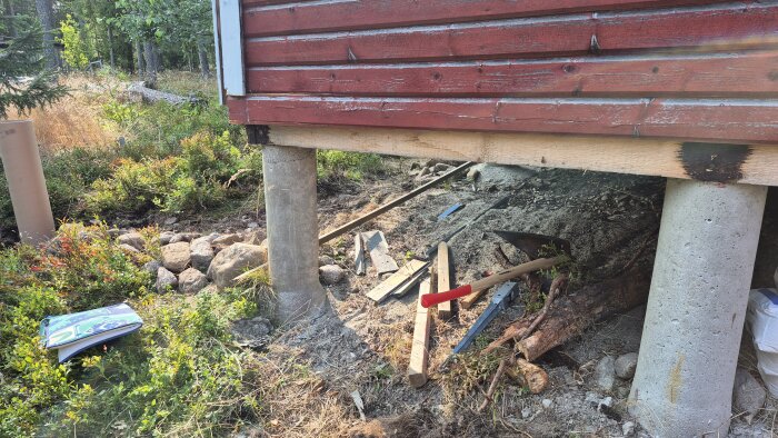 Grunden på en röd träbyggnad med betongplintar och byggmaterial, inklusive träreglar och en spade, liggande på marken.