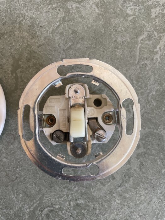 Gammal strömbrytare med grå bakgrund, centrerad metallsockel och skruvar, diskussion om byte till ny strömbrytare.