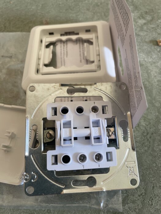 Gammal ljusströmbrytare monterad på metallplatta med skruvhål, omgiven av pappersinstruktioner och vit plastkåpa.