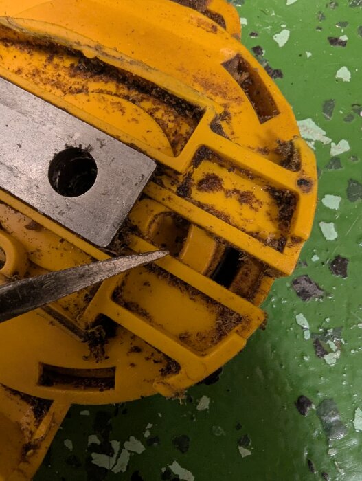 Gult verktygsfäste med spiralformad kant och metallspets som pekar på en komponent, liggande på grön yta med spår av smuts.