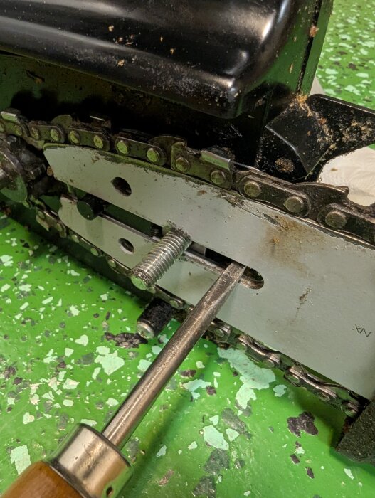 Verktyg pekar på en spiralformad kant med en skruv i ett metallsvärd på en motorsåg, grönt underlag.