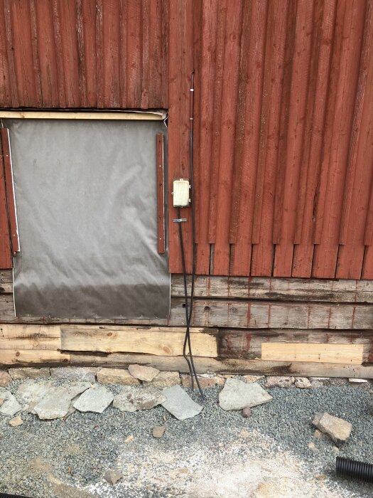 Röd träfasad under renovering med vindpapp och exponerade timmerstockar. Synlig elkabel och fiberdosa. Sten och grus vid husets grund.