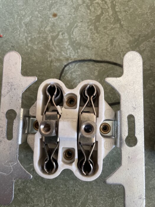 Gammalt eluttag med synliga kopplingar och skruvar, monterat på en metallram, liggande på en grön yta med en svart tråd i bakgrunden.