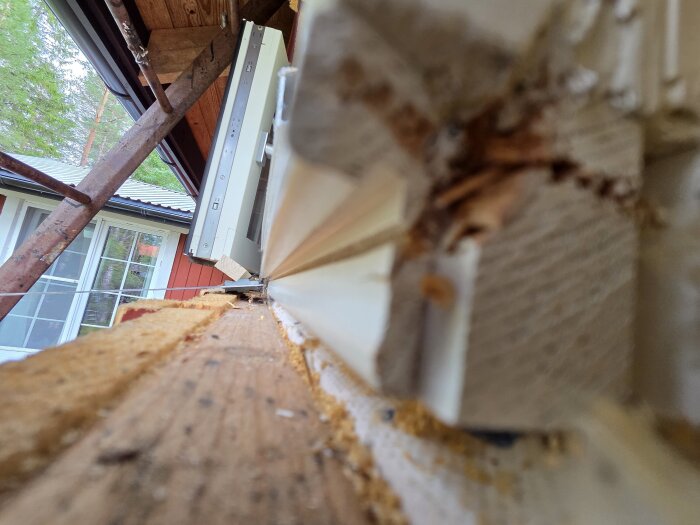 Närbild av träsida på ett hus med monterat fönsterbleck, delvis täckt av sågspån, och synligt takstöd i bakgrunden.