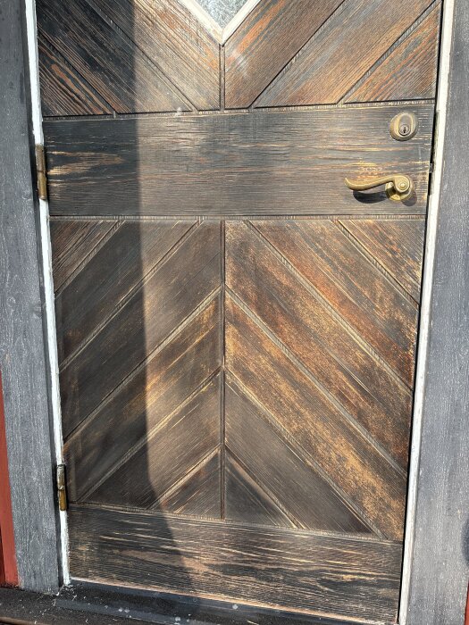 Träytterdörr med diagonalt mönster, slitage och flisor synliga. Dörrhandtag och lås i metall. Ytan har sandpapprats och ser torr ut.