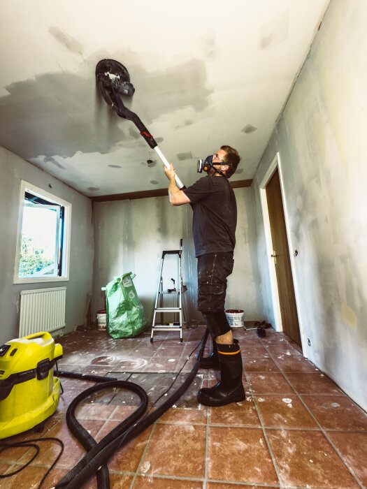 Person slipar taket med slipmaskin, bär skyddsutrustning inklusive gasmask och stövlar med tåhätta i ett rum under renovering.