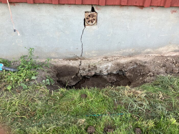 Sprucken husgrund med öppet hål och blottad jord vid en röd fasad, samt växtlighet och en bit av ett vattenrör.