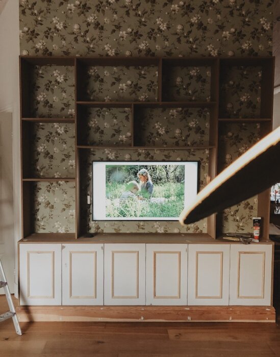Platsbyggd hylla med blommig tapet, öppna fack och TV i mitten, vita skåpdörrar nedtill; exempel på design utan synliga fästen.