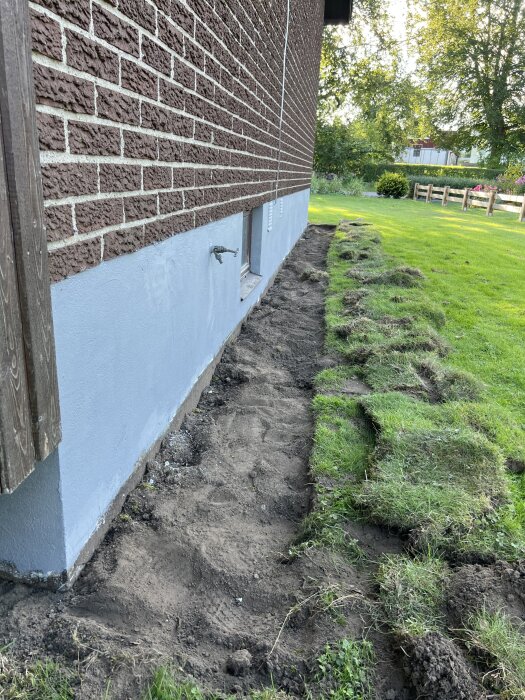 Mark längs husvägg med borttagen gräsmatta, förberedd för dränering eller planering, med synligt gräs och jord vid husets grund av tegel.