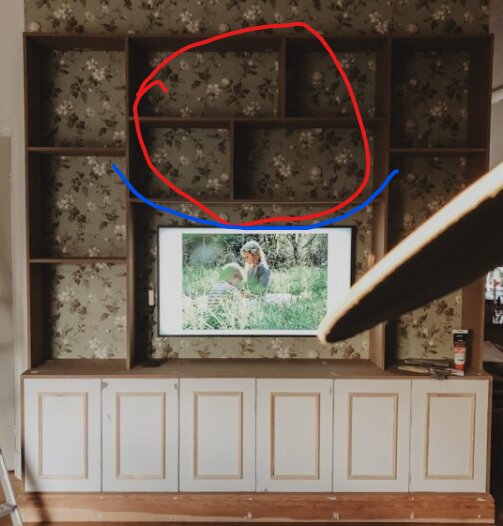 Bokhylla under uppbyggnad med inbyggd TV, omgiven av blommig tapet. Det övre mittersta hyllplanet är markerat med röd och blå markering.