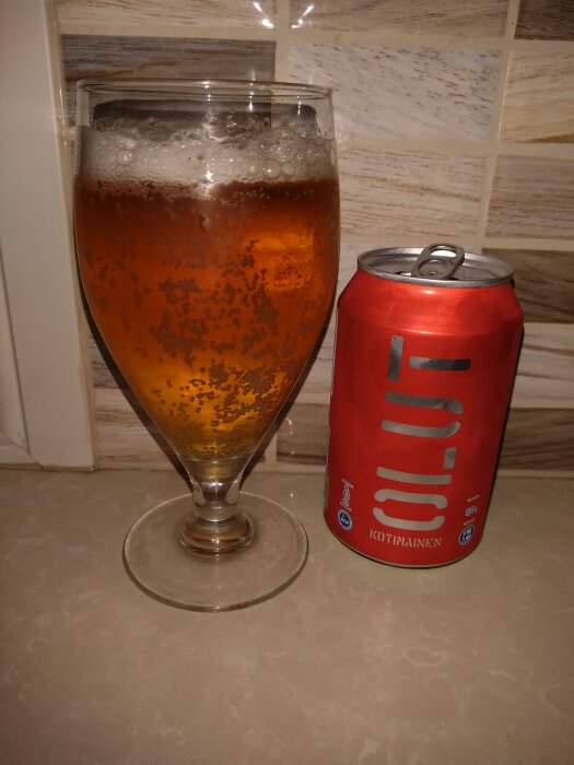 Ett glas orangetonad öl bredvid en röd ölburk med texten "OLUT" mot en kaklad bakgrund.