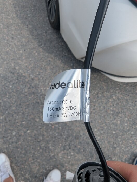 Etikett på spotlightkabel med text: "Hidealite, Art.nr.: C010, 180mA 37VDC, LED 6.7W 2700K" i en utomhusmiljö.