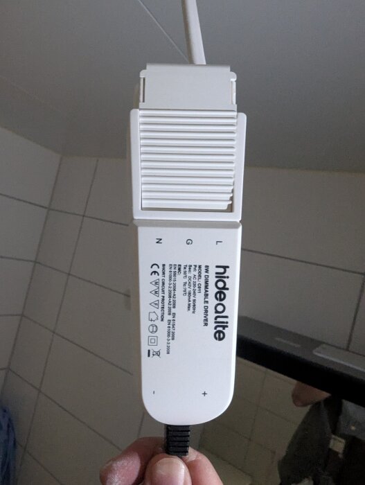 Vit Hide-a-lite spotlighttransformator i badrum, märkt "8W dimmable driver". Hållen i handen mot en kaklad vägg.