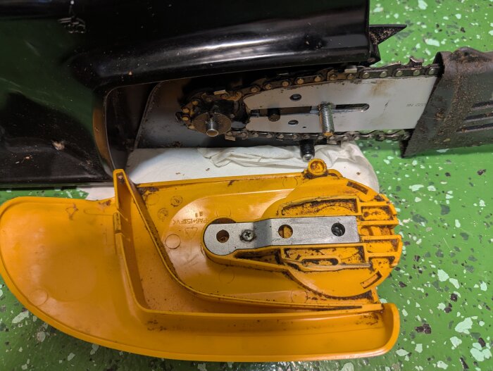 Gul kedjesågskydd med spiraldesign, visat mot framsidan av motorsågen, liggandes på ett grönt golv med små svarta och vita fläckar.