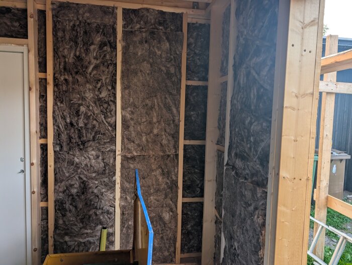 Garagets trästomme med isolering visas, det ligger fokus på väggarnas struktur och material. En dörr och några verktyg syns också.