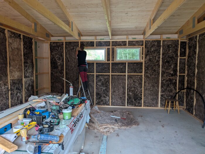 Person arbetar på garagebygge invändigt, omgiven av träreglar och isolering. Verktyg och material spridda på bänk i förgrunden.