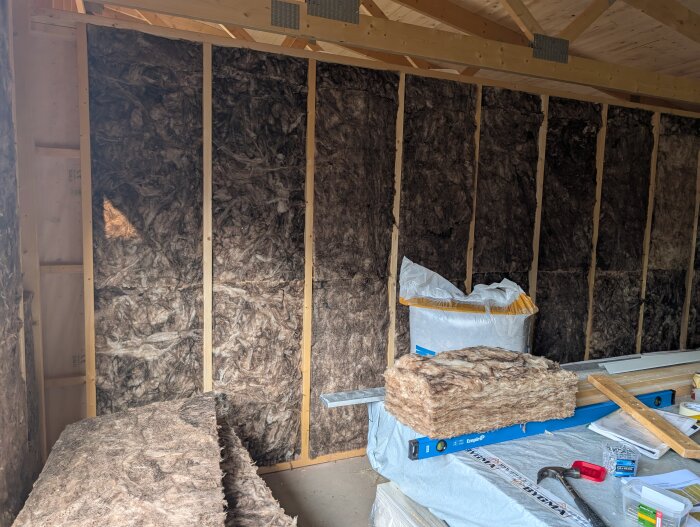 Isolerade väggar i ett garage under byggnation, med synlig trästomme och isoleringsmaterial staplat på golvet.