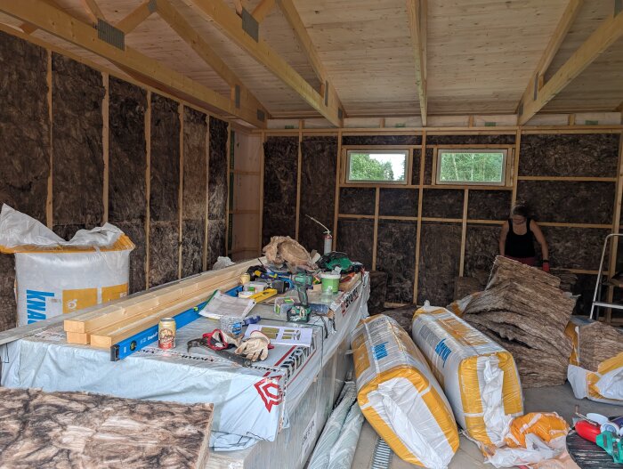 Byggarbetsplats för garage med isolering, träbjälkar och arbetsmaterial, en person arbetar med isoleringsskivor bredvid fönster.