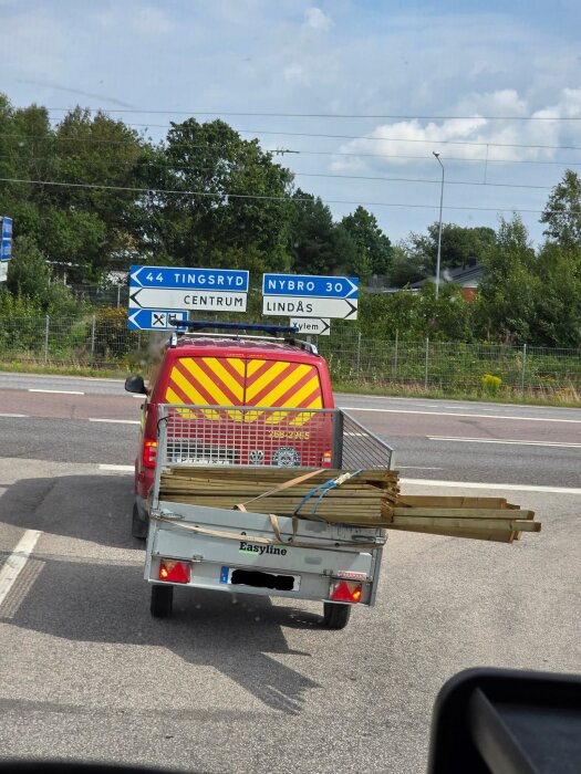 En röd bil med ett släp lastat med träbalkar, släpet verkar snett. Vägmärken visar riktning till Tingsryd och Nybro.