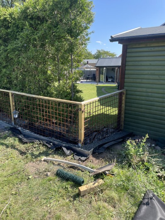 Staket och trädgård vid ett grönt hus, med nyligen avslutat staketprojekt och diverse verktyg spridda på gräset.