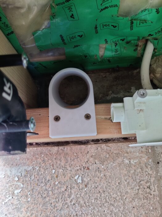 Hål i vägg för rördragning, omgiven av isolering och trä. En plastkomponent med skruvar för rörmontering är monterad mot en trälist.