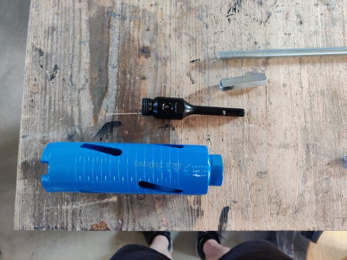 Blå hålsåg, svart adapter, gängad stång och skarvmutter på en arbetsbänk i trä.