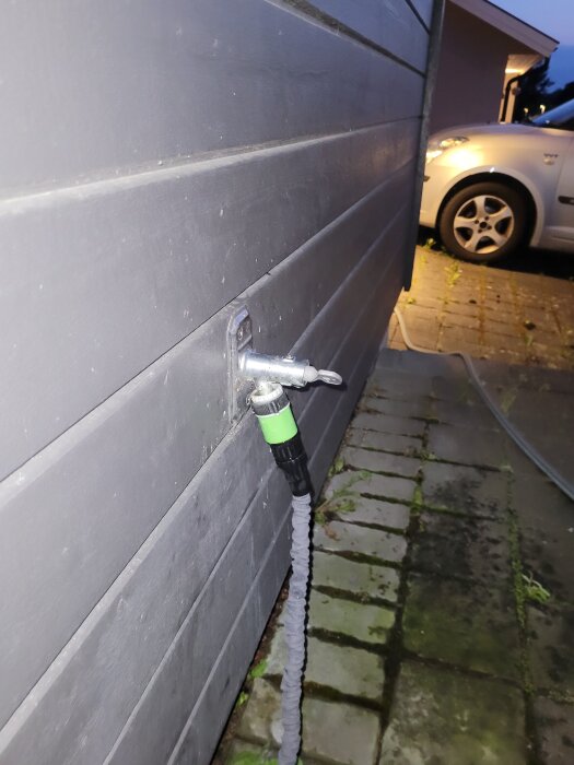 Vattenutkastare monterad på en husfasad, ansluten till en grön och svart trädgårdsslang. Stenbelagd yta syns i bakgrunden.