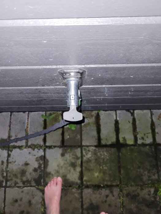 Vattenutkastare monterad på vägg, med tillhörande kran och slang, över plattbelagd yta med bar fot i bildens nederkant.