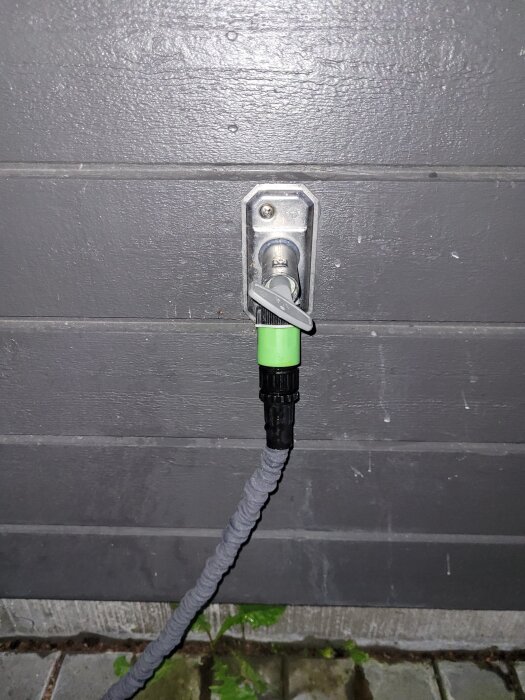 Vattenutkastare på en grå vägg med en ansluten trädgårdsslang. Ventilen är delvis öppen och omsluten av en grön och svart koppling.