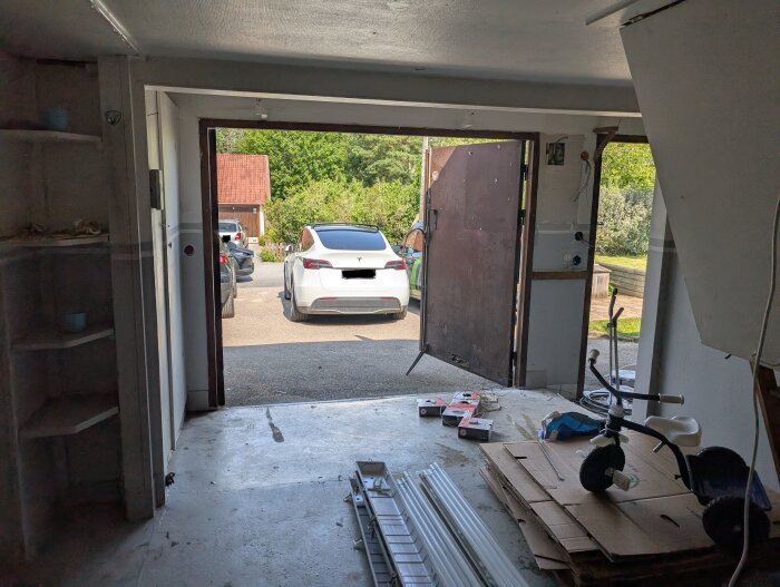 Tomt garage med öppna dörrar mot uppfart, bilar utanför, tomma hyllor och diverse skräp på golvet.