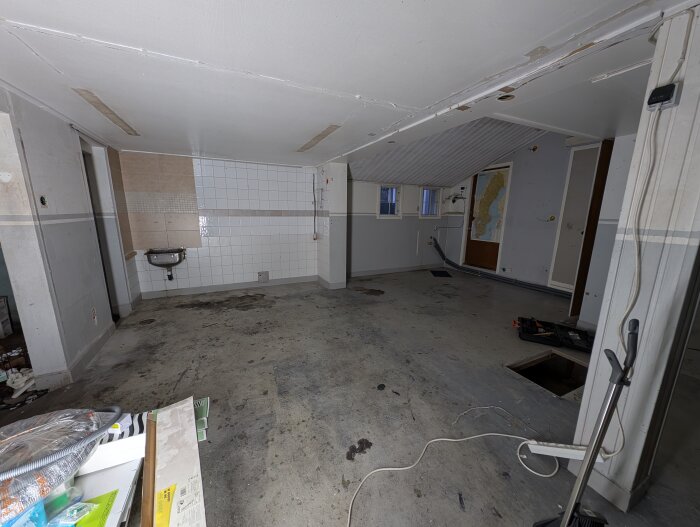 Bild på ett garage med kaklade väggar, ett handfat och ett fönster. Golvet visar tecken på tidigare arbete med betong. Verktyg och material syns i förgrunden.