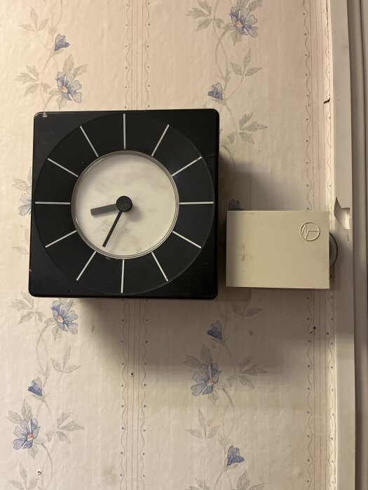 Svart fyrkantig väggklocka på blommig tapet med ett litet fyrkantigt objekt bredvid.